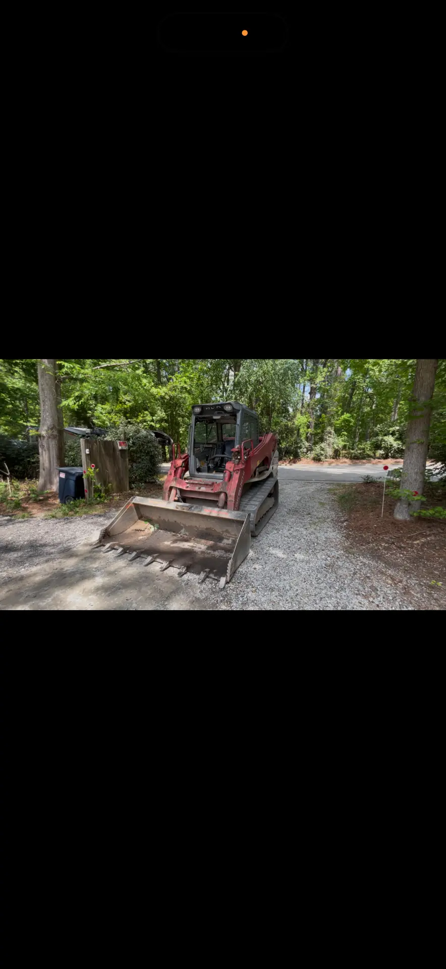 Red compact track loader parked on a gravel driveway with a bucket attachment in a wooded residential setting